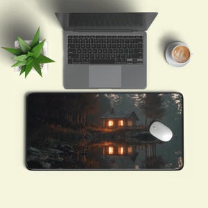 Op de afbeelding: Een computermuis en laptop liggen op een zwart rechthoekig muismat. De muismat toont een schilderachtig beeld van een hut aan een meer, omringd door bomen. Een kleine potplant en een kopje koffie zijn ook zichtbaar.