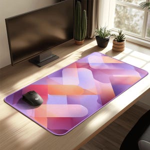 May include: A rectangular computer mouse pad with a geometric design in shades of purple, pink, and orange. A black computer mouse sits on the pad. The pad is on a wooden desk near a computer monitor.
