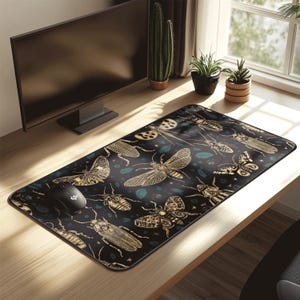 Pode incluir: Um tapete de rato retangular preto com um padrão de insetos dourados e folhas verde-azuladas. Os insetos incluem borboletas, besouros e outros desenhos detalhados. Um rato de computador preto está no lado esquerdo.