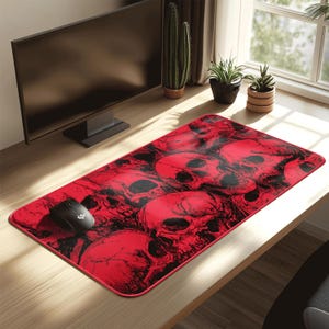 May include: A rectangular red mouse mat with a repeating pattern of black skulls. A black computer mouse is on the left side of the mat. The mouse mat is on a wooden desk, with a computer monitor and plants in the background.