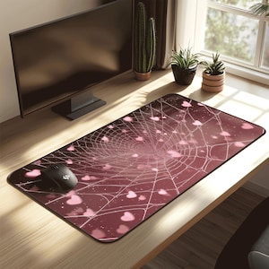 May include: A burgundy rectangular mouse mat featuring a spider web design with pink heart shapes. A black computer mouse sits on the left. The mat rests on a wooden desk, with a computer monitor in the background.