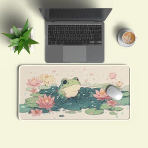 Puede incluir: Alfombrilla de ratón rectangular con una rana de dibujos animados sobre nenúfares y flores rosas y amarillas. Un ratón blanco está a la derecha. También se ven un portátil, una taza de café y una pequeña planta en maceta.