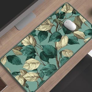 Può includere: Un tappetino da scrivania rettangolare con un design botanico di foglie e rami verdi e color crema su sfondo turchese. Un mouse per computer bianco è posizionato sul tappetino. Sono visibili anche una tazza di caffè e un monitor del computer.