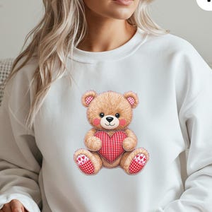 May include: White sweatshirt featuring a watercolor illustration of a teddy bear holding a red heart. The teddy bear has brown fur, pink accents, and a red gingham heart. The text "WHITE" is in the upper right corner.