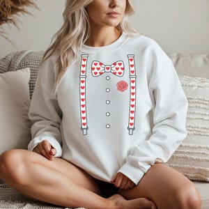 May include: White sweatshirt with a Valentine's Day design. The design includes a red heart-patterned bow tie, suspenders with red hearts, a rose, and a button-down shirt graphic.
