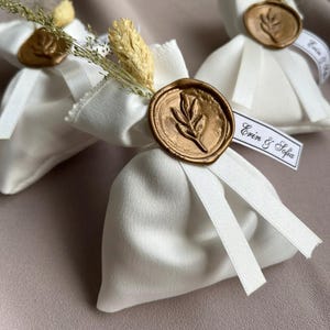 May include: Three white satin favour bags tied with white ribbons and gold wax seals. Each bag features dried flowers and a ribbon printed with "Erin & Sofia". A delicate, elegant design.