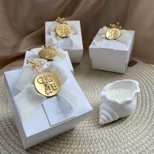 Puede incluir: Cajas de regalo blancas atadas con cintas color marfil y sellos dorados, adornadas con flores secas. También hay un candelabro blanco en forma de concha con una vela encendida. Las cajas tienen una etiqueta con nombres y una fecha.