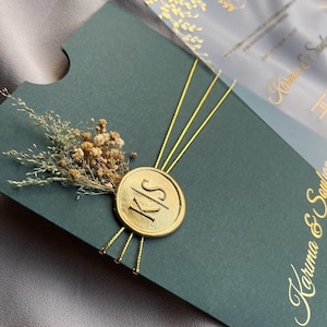 Może przedstawiać: Ciemnozielone zaproszenie ślubne ze złotymi akcentami. Zaproszenie posiada złotą pieczęć z inicjałami "K/S" i jest ozdobione suszonymi kwiatami. Widoczna jest przezroczysta nakładka ze złotymi wzorami kwiatowymi.