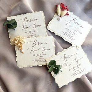 Op de afbeelding: Vier crèmekleurige kaarten met een getextureerde afwerking en gescheurde randen, elk versierd met bloemaccenten. De kaarten tonen elegante kalligrafie en de tekst "Berre nin Kina Geesi", een datum en een tijd. De kaarten zijn gerangschikt op een neutrale stof.