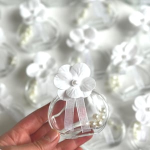 Puede incluir: Pequeños frascos de vidrio transparente decorados con flores de papel blanco, perlas y cinta blanca. Los frascos redondos están llenos de perlas. La flor tiene un centro de perlas. Ideal para recuerdos de boda.