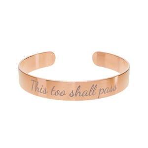 Puede incluir: Pulsera de brazalete de oro rosa con el texto grabado "This too shall pass". La pulsera abierta tiene un acabado liso y pulido y un diseño simple y elegante. Una pieza de joyería motivacional.