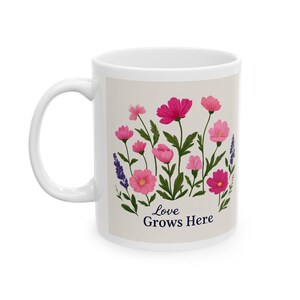 Op de afbeelding: Witte keramische mok met een bloemenontwerp. Het ontwerp toont roze en paarse bloemen en groene bladeren. De mok heeft de tekst "Love Grows Here" in donkerblauw. De mok heeft een wit handvat.