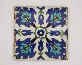 May include: Square ceramic tile with a floral design. The tile has a white background with a symmetrical pattern of blue and turquoise flowers and green accents. The edges have a distressed, aged appearance.