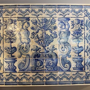 Peut inclure: Une œuvre d'art murale rectangulaire composée de plusieurs carreaux carrés. Chaque carreau présente un motif bleu et blanc avec des chérubins, des motifs floraux et des éléments décoratifs. L'esthétique générale rappelle l'art du carrelage céramique classique.
