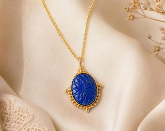 Könnte beinhalten: Eine goldene Halskette mit einem ovalen Anhänger aus tiefblauem Lapislazuli, der mit einem Blumenmuster graviert ist. Der Anhänger ist von einem perlenartigen Goldrand umgeben und hängt an einer feinen Goldkette, was ein edles Bild ergibt.