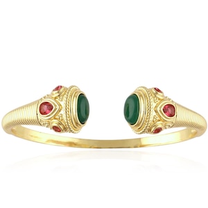 May include: A gold-tone open cuff bracelet featuring a ribbed band design. Each end is adorned with a central oval green stone surrounded by smaller, teardrop-shaped red stones set within intricate, textured gold-tone metal detailing.