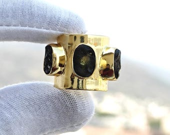 May include: A gold ring with four dark green, rough-cut stones. The ring has a geometric design, with the stones set into the gold band. The ring is held by a gloved hand.