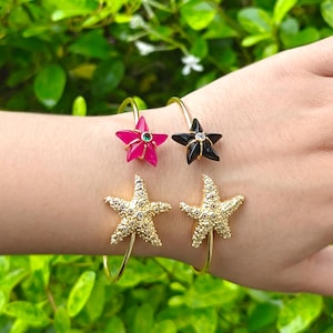 May include: Two gold-tone cuff bracelets featuring starfish designs. One bracelet has a bright pink starfish with a small green center stone, and the other has a black starfish with a clear center stone. Two larger gold starfish with clear stones.
