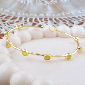 K&ouml;nnte beinhalten: Ein goldfarbenes Armreif-Armband mit sechs gleichm&auml;&szlig;ig verteilten, hellgr&uuml;nen Edelsteinen. Das offene Armband hat eine kleine Schlaufe an einem Ende. Es liegt auf einer rosa Oberfl&auml;che.