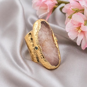 May include: A gold ring featuring a large, pale pink stone. The ring has a textured band and is embellished with small green gemstones. The background includes soft pink flowers and a silky, grey fabric.