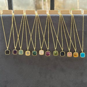Puede incluir: Una colección de collares dorados, cada uno con un colgante cuadrado con una piedra preciosa de diferente color. Las piedras preciosas incluyen negro, morado, verde y otros colores. Los collares se exhiben en un soporte de madera.