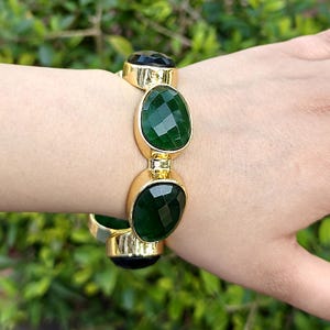 May include: Gold-toned bracelet featuring three large, faceted, oval-shaped green gemstones and two smaller black gemstones. The bracelet is shown on a wrist against a blurred green background.