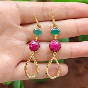 May include: A pair of gold-tone drop earrings featuring a small oval green stone at the top, a larger round faceted magenta stone in the center, and an irregular open-form gold-tone metal loop at the bottom, suspended from a hook-style wire.