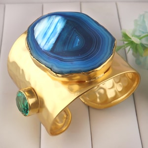 May include: A gold-toned cuff bracelet featuring a large, blue agate stone with concentric patterns. The bracelet has a hammered texture and a smaller, green gemstone accent. A stylish piece of jewelry.