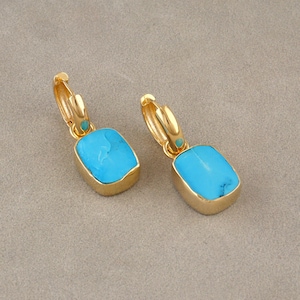 May include: A pair of gold-colored hoop earrings with rectangular, light blue turquoise stones. The earrings have a small turquoise accent near the hoop closure. The background is a neutral, light brown color.