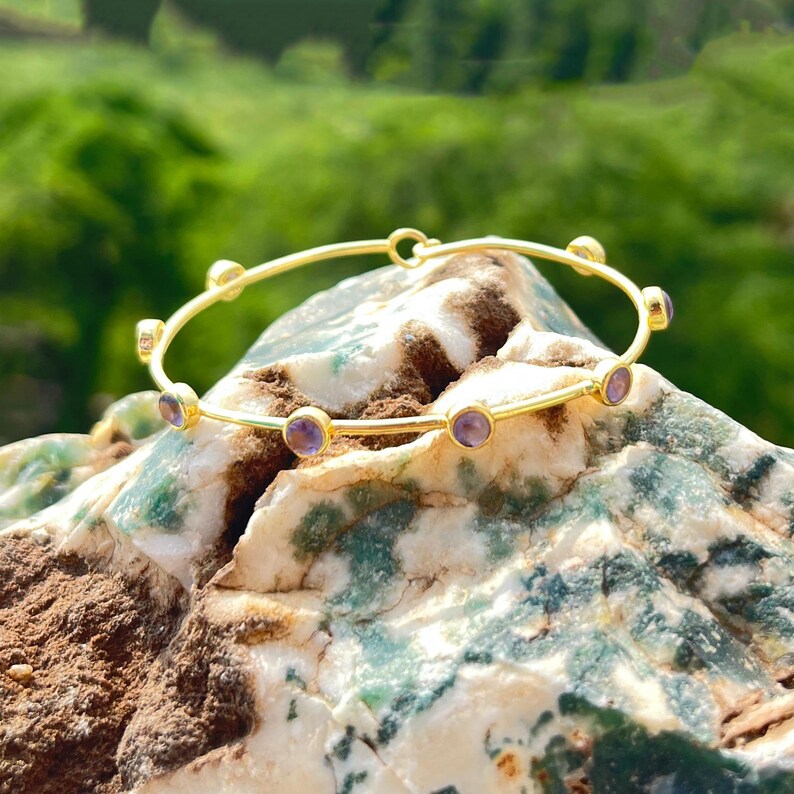 K&ouml;nnte beinhalten: Ein goldfarbenes Armreif-Armband mit kleinen, runden, lila Edelsteinen, die gleichm&auml;&szlig;ig &uuml;ber das Band verteilt sind. Das Armband liegt auf einer strukturierten, mehrfarbigen Felsformation mit verschwommenem gr&uuml;nem Hintergrund.