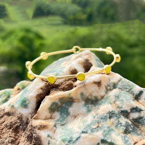 K&ouml;nnte beinhalten: Ein goldfarbenes Armreif-Armband mit kleinen, runden, gelben Edelsteinen, die gleichm&auml;&szlig;ig &uuml;ber das Band verteilt sind. Das Armband wird auf einer strukturierten, mehrfarbigen Felsformation mit verschwommenem gr&uuml;nem Hintergrund pr&auml;sentiert.