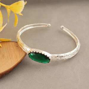 May include: A silver-toned open cuff bracelet featuring a textured, bark-like surface pattern and a single, large, oval-shaped faceted emerald green gemstone set in a scalloped bezel at the center of the band.