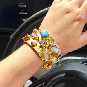 May include: A wide gold-tone cuff bracelet with a hammered texture, set with various gemstones. The bracelet features turquoise, pink, and white stones. The bracelet is worn on a wrist.