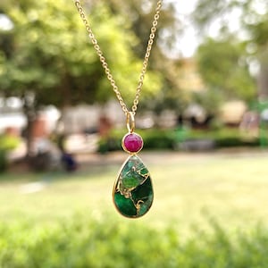 May include: A gold-colored necklace with a pendant featuring a teardrop-shaped green stone with gold veining, topped by a small, round, red stone. The necklace hangs against a blurred green background.