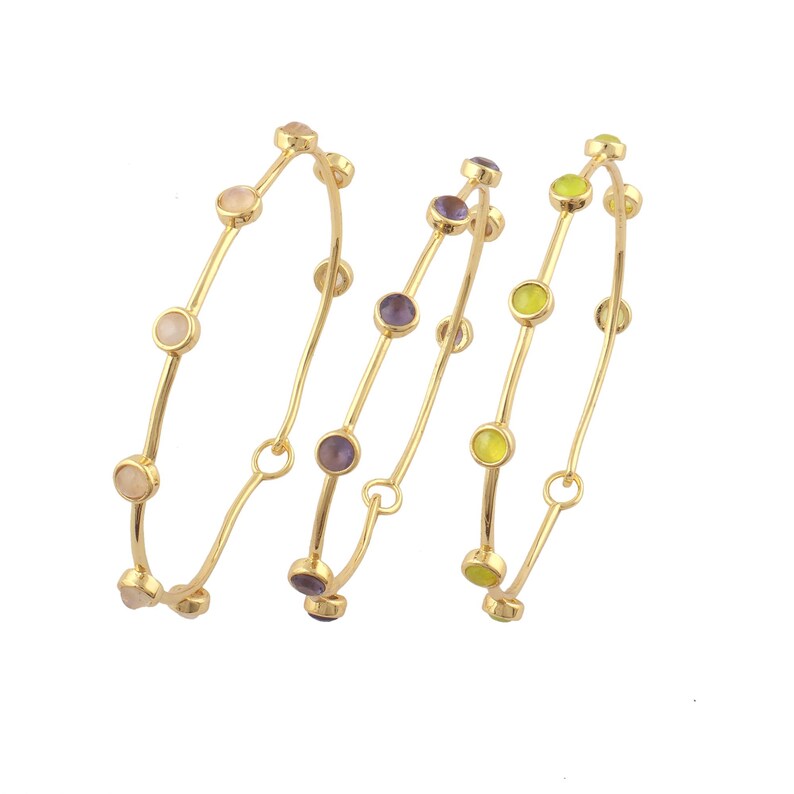 K&ouml;nnte beinhalten: Drei goldfarbene Armreifen mit bunten Edelsteinen. Die Armreifen haben ein zartes Design, wobei jeder eine Reihe runder Edelsteine in den Farbt&ouml;nen Rosa, Lila und Gelb aufweist. Die Armreifen werden vor einem wei&szlig;en Hintergrund pr&auml;sentiert.