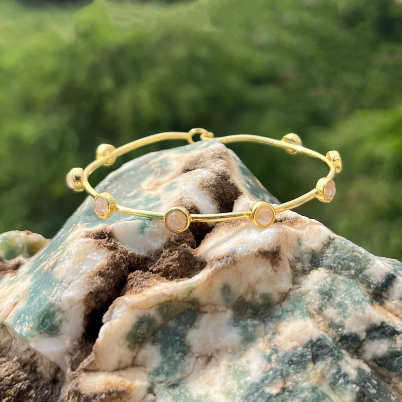 K&ouml;nnte beinhalten: Ein goldfarbenes Armreif-Armband mit sechs runden, hellrosa Edelsteinen, die gleichm&auml;&szlig;ig &uuml;ber das Band verteilt sind. Das Armband wird auf einem strukturierten, mehrfarbigen Felsen mit verschwommenem gr&uuml;nem Hintergrund pr&auml;sentiert.