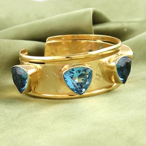 Può includere: Un bracciale a polsino dorato con tre pietre preziose triangolari blu chiaro. Il bracciale ha una fascia larga con un design curvo ed è posto su uno sfondo verde. Le pietre preziose hanno un aspetto sfaccettato.