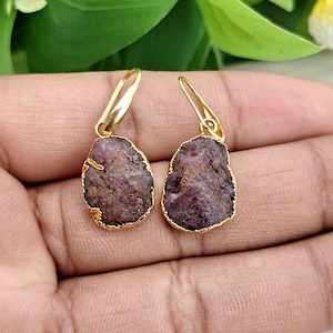 May include: A pair of gold-colored earrings with a natural, rough-cut, dark purple gemstone. The gemstones are teardrop-shaped and have a gold-colored border. The earrings are displayed against a neutral background.