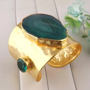 May include: A wide gold-tone cuff bracelet featuring a large, polished, dark green agate slice with natural banding and a smaller, round, faceted emerald-green gemstone set in a gold-tone bezel on the side of the hammered metal band.