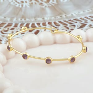 K&ouml;nnte beinhalten: Ein goldfarbenes Armreif-Armband mit sechs runden, lila Edelsteinen, die gleichm&auml;&szlig;ig &uuml;ber das Band verteilt sind. Das Armband liegt auf einer hellrosa Oberfl&auml;che, mit einem verschwommenen, dekorativen Hintergrund.