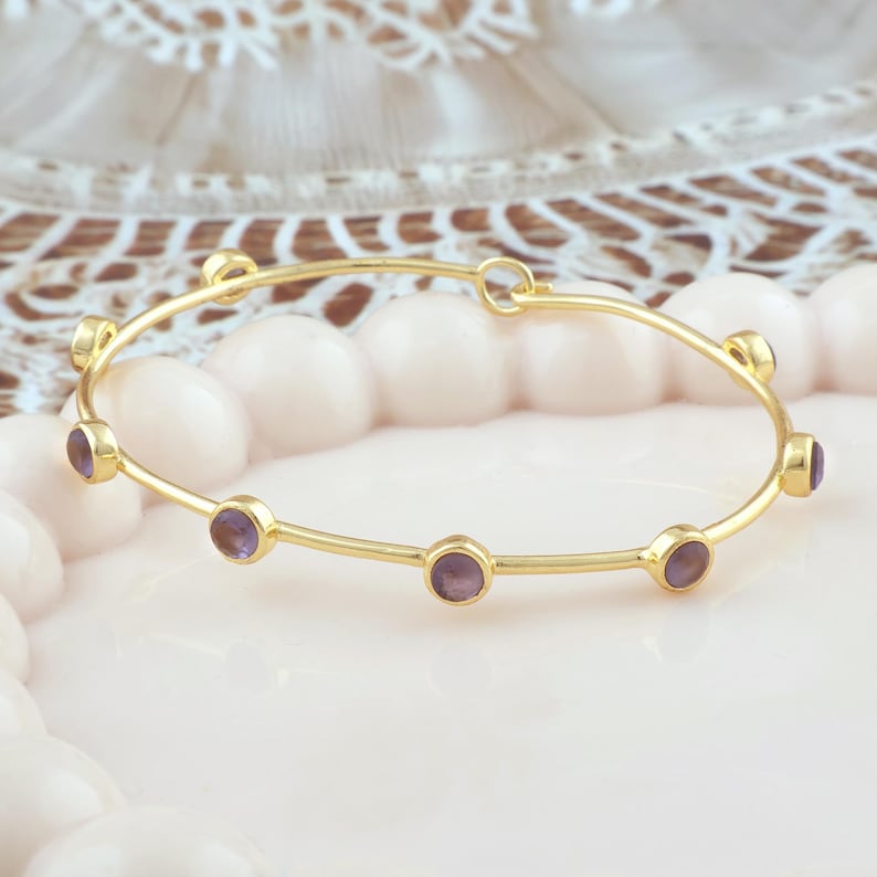 K&ouml;nnte beinhalten: Ein goldfarbenes Armreif-Armband mit sechs runden, lila Edelsteinen, die gleichm&auml;&szlig;ig &uuml;ber das Band verteilt sind. Das Armband liegt auf einer hellrosa Oberfl&auml;che, mit einem verschwommenen, dekorativen Hintergrund.