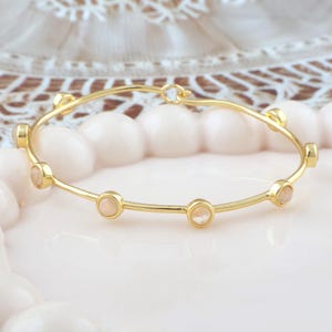 K&ouml;nnte beinhalten: Ein goldfarbenes Armreif-Armband mit sechs kleinen, runden, hellpfirsichfarbenen Steinen, die gleichm&auml;&szlig;ig &uuml;ber das Band verteilt sind. Das Armband hat ein schlichtes, elegantes Design. Der Hintergrund ist ein weiches, verschwommenes Rosa und Wei&szlig;.