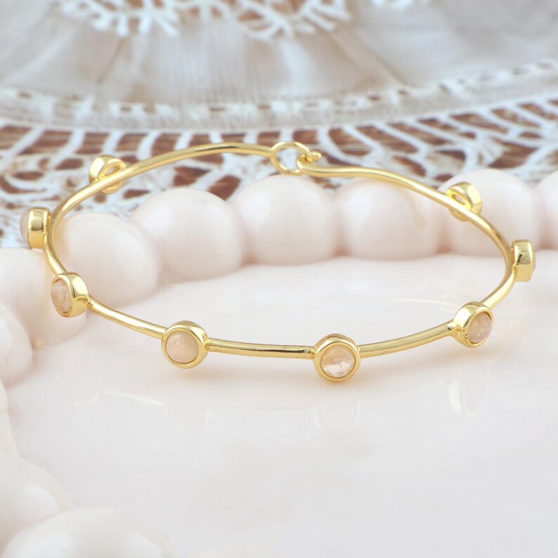 K&ouml;nnte beinhalten: Ein goldfarbenes Armreif-Armband mit sechs kleinen, runden, hellpfirsichfarbenen Steinen, die gleichm&auml;&szlig;ig &uuml;ber das Band verteilt sind. Das Armband hat ein schlichtes, elegantes Design. Der Hintergrund ist ein weiches, verschwommenes Rosa und Wei&szlig;.