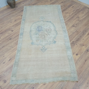 Puede incluir: Una alfombra rectangular vintage con un diseño de medallón central en tonos azules y beige. La alfombra tiene un aspecto descolorido, con un borde en un tono azul más claro. La alfombra está colocada sobre un suelo de madera.