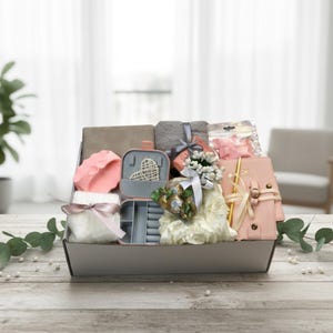 May include: A gift basket containing a selection of items: a pink jewellery box, a pink notebook with a gold pen, a grey towel, and a white faux fur item tied with a pink ribbon. The basket is on a wooden surface.