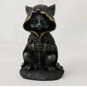 May include: A black cat figurine wearing a hooded cloak with gold trim, holding a staff. The cat has bright green eyes and is sitting on a textured base. The figurine is a decorative item.