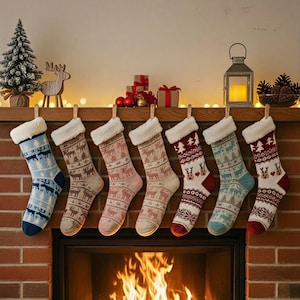 May include: Seven Christmas stockings with reindeer and snowflake patterns hang above a fireplace with a burning fire. The stockings are in various colors, including blue, brown, and red, with white faux fur cuffs. A small Christmas tree and other decorations are on the mantel.