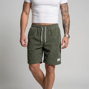 Puede incluir: Pantalones cortos de pana verde oliva con cintura con cordón blanco. Los pantalones cortos tienen una pequeña etiqueta blanca en la parte delantera. El modelo lleva una camiseta blanca. Los pantalones cortos son hasta la rodilla.