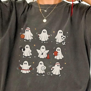May include: A charcoal gray short-sleeve t-shirt featuring a graphic print of nine white ghosts engaged in various sports activities, including basketball, soccer, weightlifting, and hula hooping, accented with small gold stars scattered throughout.