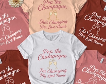 Puede incluir: Una colección de cinco camisetas de manga corta en colores melocotón, blanco, óxido y rosa empolvado. Cada camiseta incluye el texto Pop the Champagne, un gráfico de dos copas brindando y la frase She's Changing Her Last Name o I'm Changing My Last Name.